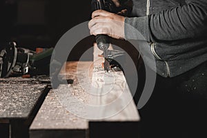 A man works with a drill in his workshop. Carpenter drills with a hand power tool in a dark room