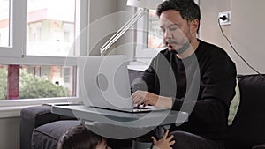 A man works on his laptop at home on the couch, while his one-year-old child holds on to the table