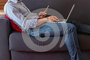 man works on his laptop and looks at the smartphone lying on the sofa