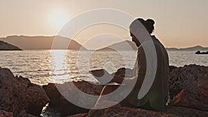 A man works on a laptop while sitting on a stone by the sea at sunrise.