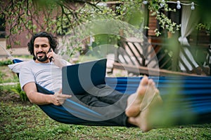 A man works with a laptop and smartphone in a hammock in a country house