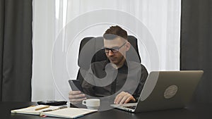 Man works in modern office. Young successful businessman in glasses looking on phone sitting at his office desk. Male broker check