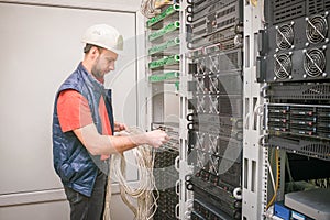 A man works in the server room of a data center. A technician connects a cable to a stack of managed switches. An engineer in a