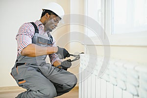 Man in workwear overalls using tools while installing or repairing heating radiator in room
