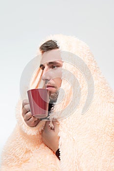 The man is wrapped in a blanket, holding a mug with a drink in his hands, the man is sick with the flu.