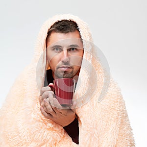 The man is wrapped in a blanket, holding a mug with a drink in his hands, the man is sick with the flu.