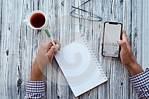 Man writes by his left hand in a notebook while holding a phone. Glasess and cup of tea. Conceptual photo of lefthanded