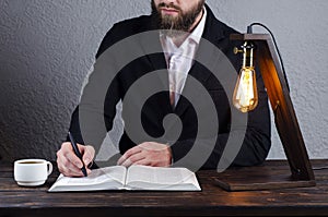 The man writes in a notebook. Open book Bible. Man is sitting at the table. Book reading. Desktop. Edison`s table lamp.