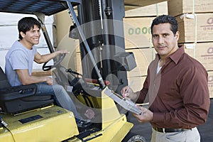 Man Writing On A Clipboard With Worker Driving Forktruck At Warehouse