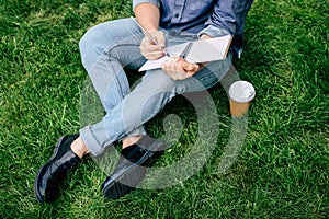 Man writing on notebook and drinking coffee from paper cup while sitting on green grass