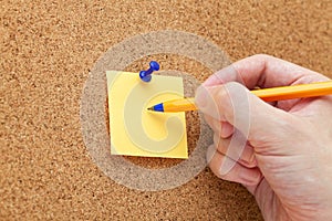 A man writing on a paper note with a ballpoint pen that is pinned on a cork board