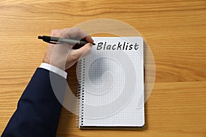 Man writing word Blacklist in notebook at wooden table, top view