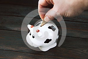 A man& x27;s hand puts a coin in the piggy bank on a wooden background.Saving money. Family budget. Money box on a wooden photo