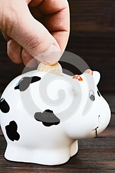 A man& x27;s hand puts a coin in the piggy bank on a wooden background.Saving money. Family budget. Money box on a wooden photo