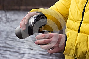 A man in a yellow jacket is pouring tea from a thermos into a cup. Cool spring day near the lake in the forest