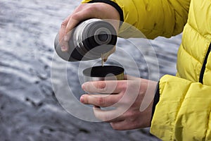 A man in a yellow jacket is pouring tea from a thermos into a cup. Cool spring day near the lake in the forest