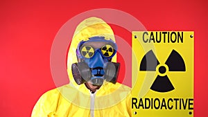 Man in Yellow Protective Suit, Protective Mask with Poster Caution radiation shakes its head negatively. Behind the man