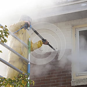 Man in yellow rain suit cleans paint from brick wall of house fa
