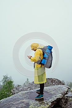 A man in a yellow raincoat with a backpack on his back stands on a rock in rainy weather against a foggy landscape and uses a