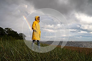 Man in yellow raincoat with hood walk on the beach in rainy weather, looks at dramatic cloudy sky and sea, rainbow on background