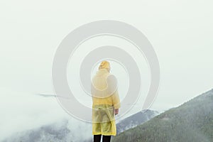 A man in a yellow raincoat stands on top of a mountain in rainy weather against a foggy landscape, view from the back