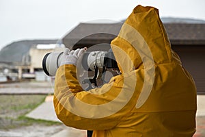 A man in a yellow raincoat takes photos with a long-focus lens fixed on a monopod tripod. Rear view