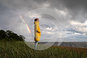 Man in yellow raincoat wear red hat standing on the beach in rainy weather, looks at dramatic cloudy sky and sea, rainbow on