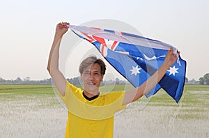 The man in yellow shirt is holding Australia flag in his hands and raising to the end of the arm at the back on nature view
