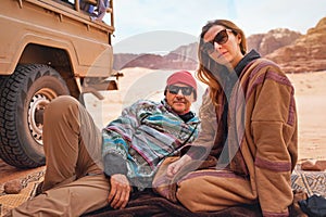 Man and younger woman wearing traditional Bedouin warm coat - bisht -  sitting or laying on ground blanket near off road vehicle