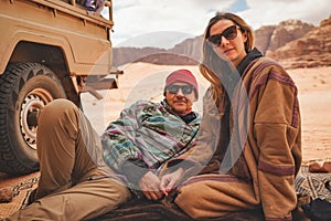 Man and younger woman wearing traditional Bedouin warm coat - bisht - sitting or laying on ground blanket near off road vehicle photo