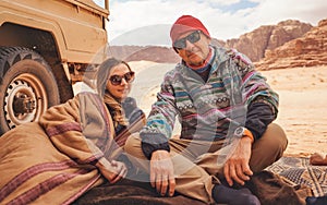 Man and younger woman wearing traditional Bedouin warm coat - bisht - sitting or laying on ground blanket near off road vehicle photo