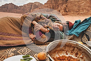 Man and younger woman wearing traditional Bedouin warm coat - bisht - resting, lying down on ground blanket near off road vehicle photo