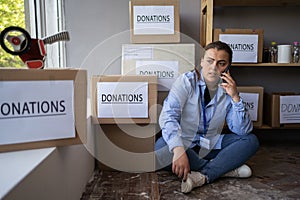 Manager of charity center sitting near boxes with donations lettering and smartphone while working in charity center