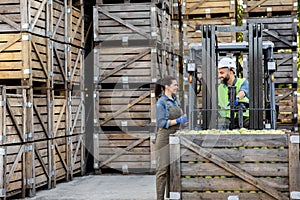 Manager and driver work in warehouse with many wooden boxes, load boxes of fruit for storage
