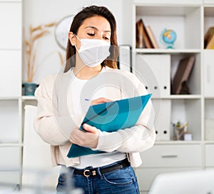 Manager girl stands in a protective mask in the office and fills out documents on a folder