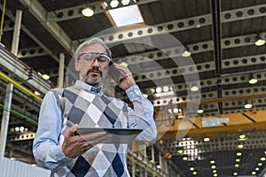 Manager Looking At Digital Tablet Screen And Talking On Smartphone In A Factory