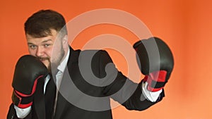 Manager in suit and gloves is boxing and throws punches at the camera in slowmo on orange background. Cheerful plump
