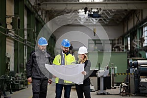 Manager supervisor, engineer and industrial worker in uniform discussing blueprints in large metal factory hall.