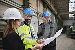 Manager supervisor, engineer and industrial worker in uniform discussing blueprints in large metal factory hall.