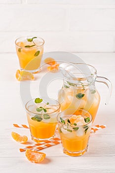 Mandarin cocktail with ice and mint in beautiful glasses and jug, fresh ripe citrus on white wooden background. Sweet orange juice