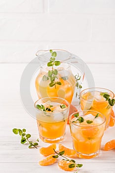 Mandarin cocktail with ice and mint in beautiful glasses and jug, fresh ripe citrus on white wooden background. Sweet orange juice