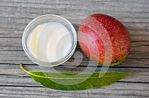 Mango body butter in a glass bowl and fresh ripe organic mango fruit and leaf on old wooden background.