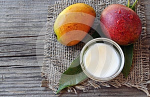 Mango body butter in a glass bowl and fresh ripe organic mango fruit and leaves on old wooden background.