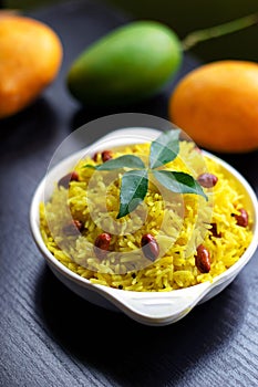 Mango rice in bowl , an Indian dish garnished with curry leaves & roasted peanut with raw ripe mangoes in the background on black