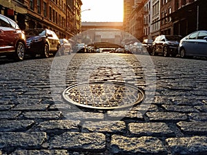 Manhole cover in an old cobblestone street lined with parked cars in the Tribeca neighborhood of New York City