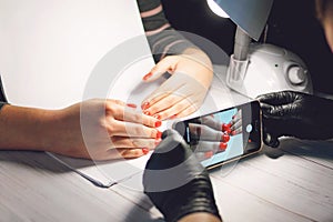 Manicure specialist in black gloves takes pictures of hands with red nails by phone. Manicurist in beauty salon. How shoot nail