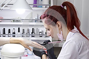 A manicurist in a beauty salon in a mask applies gel with a brush to the nails of a client.