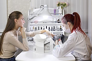 A manicurist in a beauty salon in a mask treats a client`s nails before applying the gel.