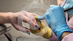 Manicurist in rubber gloves applies black shellac gel polish to the nail of a Caucasian woman's hand.