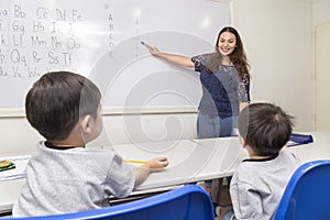 A female English teacher teaching two little boys, explaining ABC alphabet on board in classroom at school